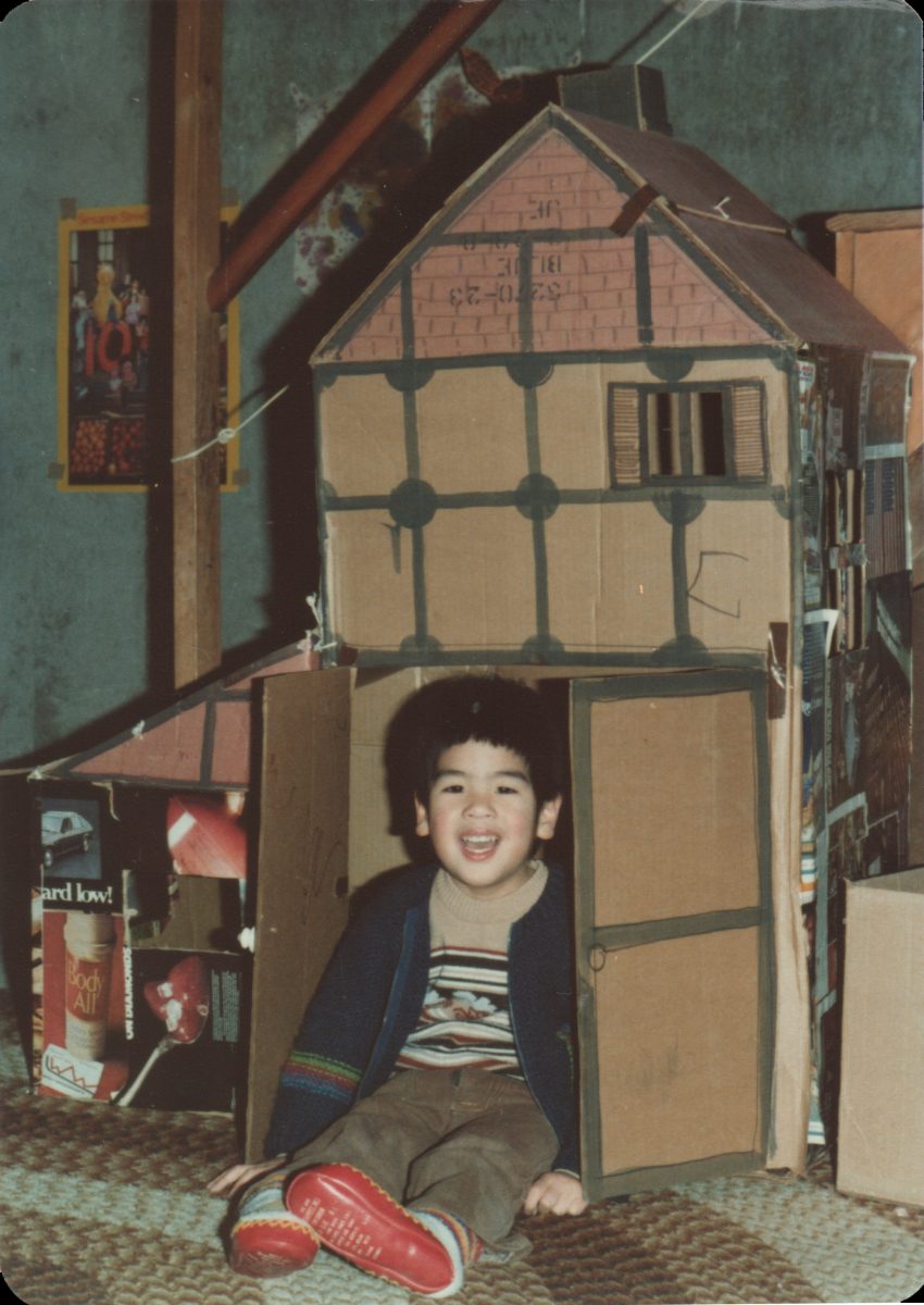 Smiling child sits in front of a homemade cardboard house with open doors, as if a fine artist has crafted a cozy scene indoors.