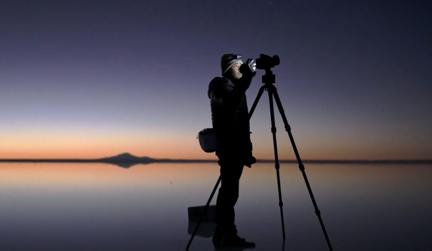 A photographer captures a stunning shot with a camera on a tripod by a calm lake at sunset, skillfully blending elements of nighttime landscape photography.