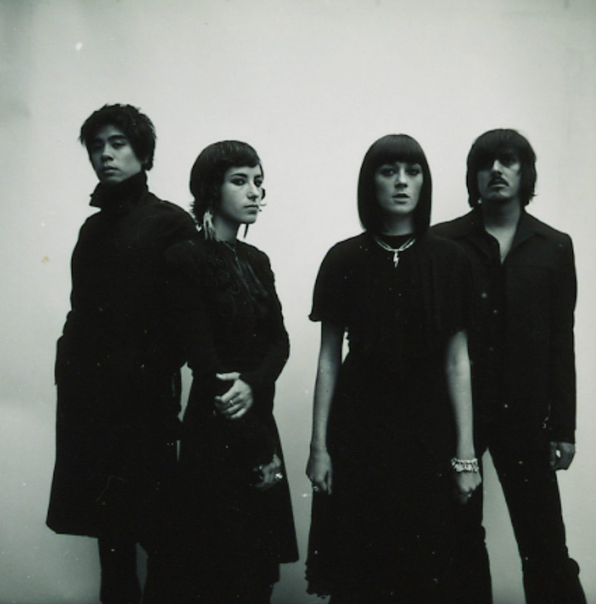 Four people in dark clothing stand side by side against a plain, light background, their serious expressions evoking the dramatic flair of drone light art.