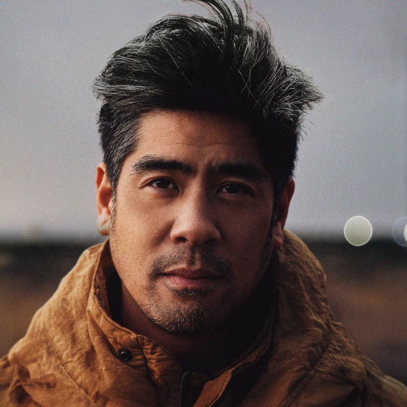 Man with dark hair and a brown jacket looks at the camera outdoors in soft, natural light—a futuristic vibe reminiscent of nighttime landscape photography.